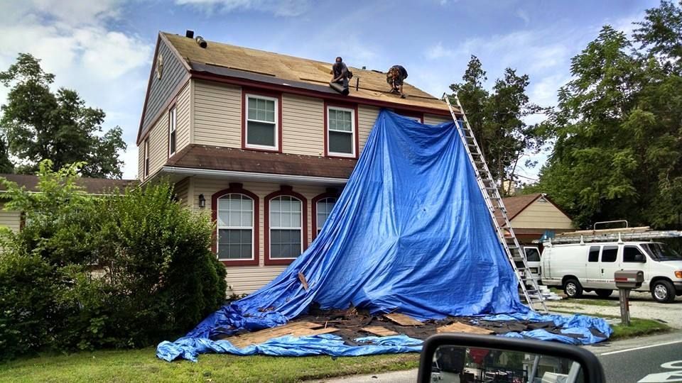 Two workers on a house roof covered with a blue tarp and a ladder.