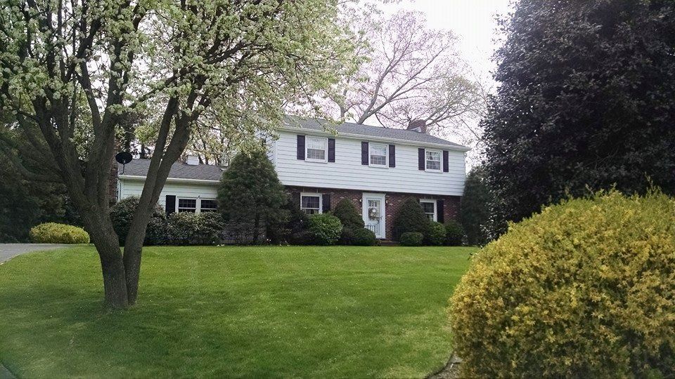 Two-story white house with black shutters, brick entrance, and a green lawn. Trees and bushes surround the house.