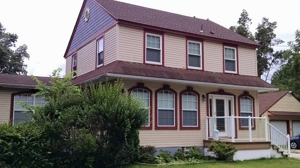 Two-story beige house with red trim, brown roof, and small porch. Bushes in front.