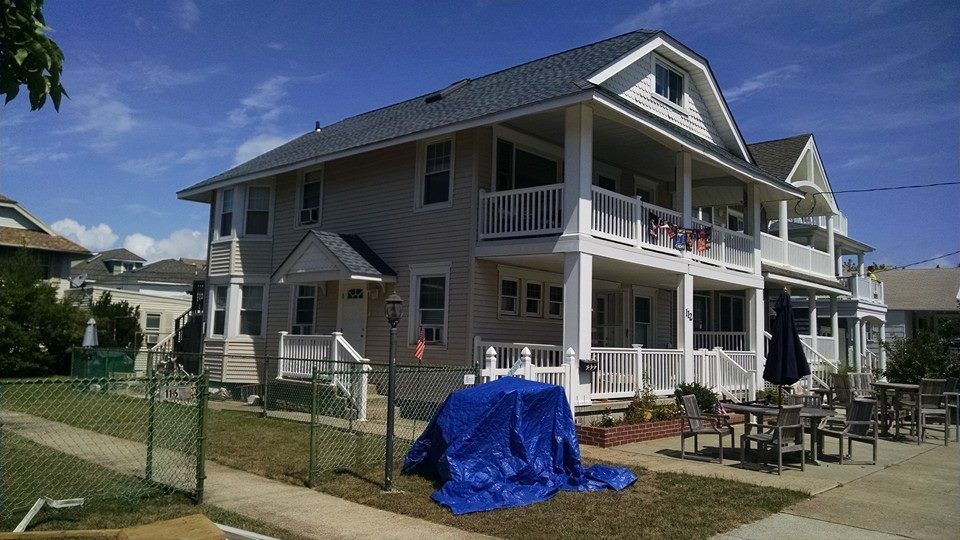 Two-story beige beach house with balconies, white railings, and a blue tarp on the front lawn under a bright blue sky.