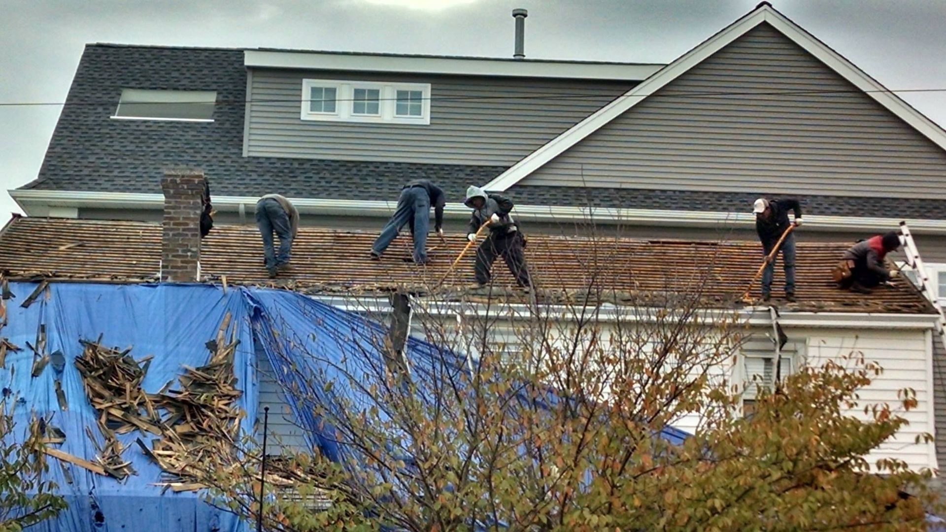 Roofers removing old shingles from a house with a gray exterior on a cloudy day. Blue tarp covers debris.