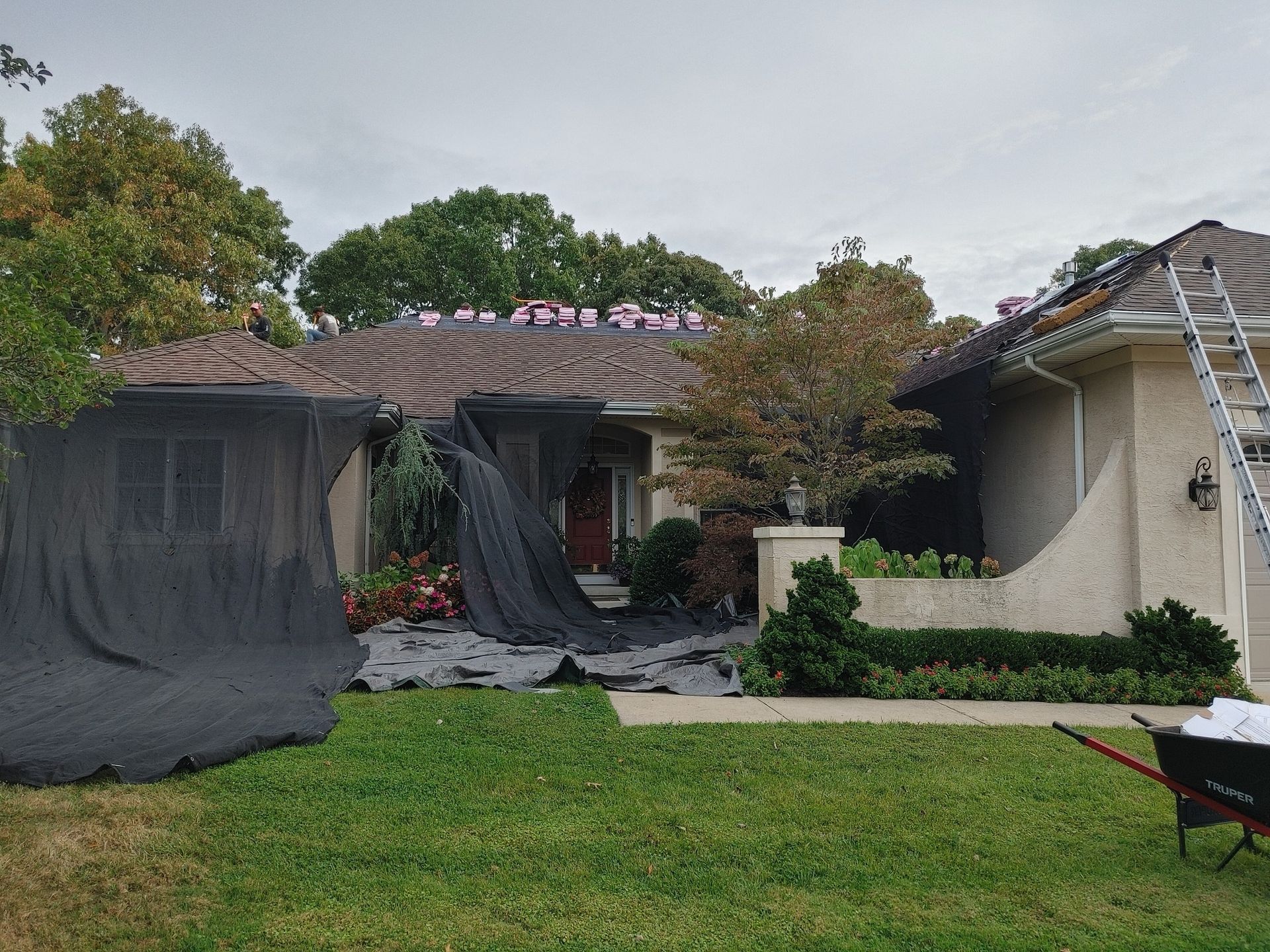 House under roof repair, covered in black netting, ladder on side. Overcast day.