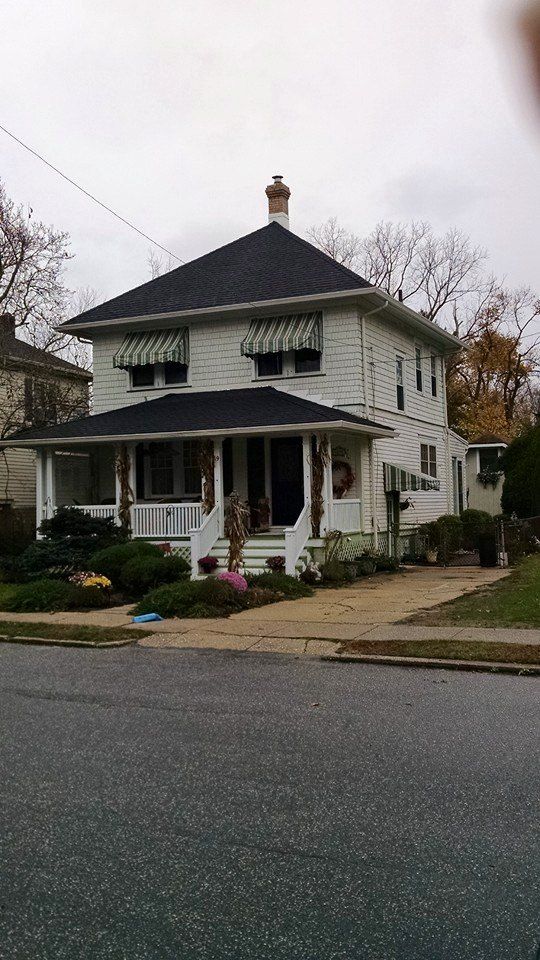 Two-story white house with black roof, front porch, awnings, and landscaped yard on a street.