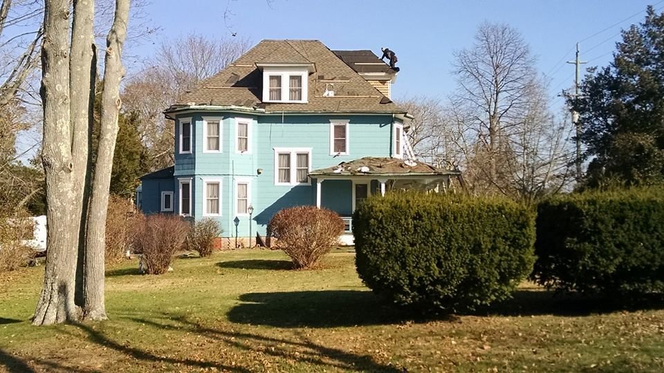 Turquoise house with dormers, a porch, and a brown roof. Green lawn with bushes and trees in front.