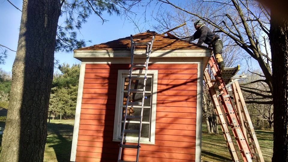 Person on a ladder, working on the roof of a small, red shed in a sunny outdoor setting.