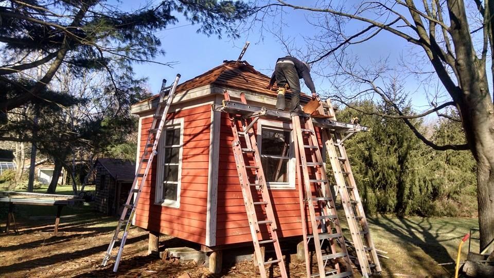 Person on a small red building's roof, using ladders. Sunny day, trees in background.