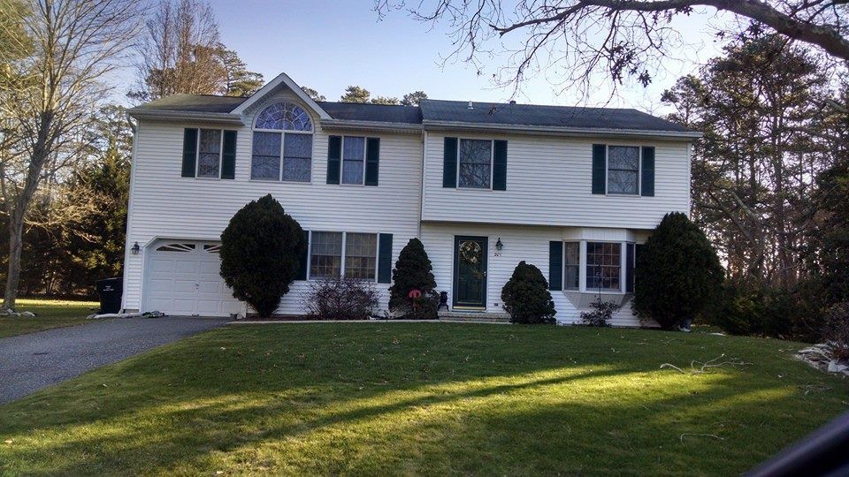 Two-story white house with green shutters and front lawn, driveway on the left, trees in the background.