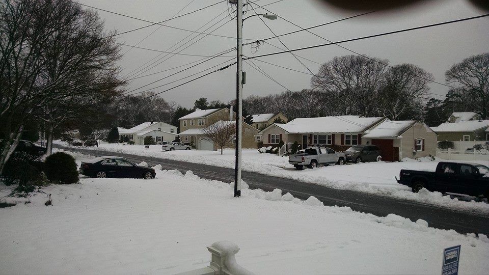 Snow-covered suburban street with houses and parked cars. Overcast sky, bare trees, and power lines.