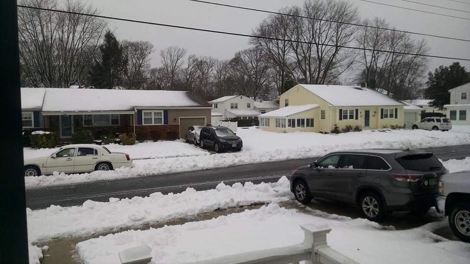 Snowy suburban street scene with houses, cars, and snow-covered lawns and sidewalks.