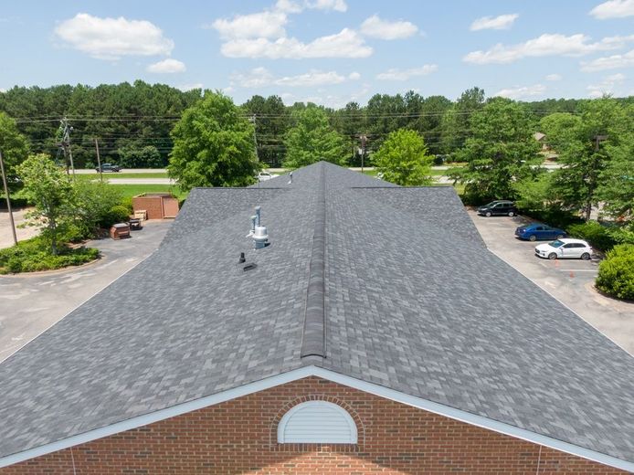 Aerial view of a gray shingled roof on a brick building, surrounded by trees and a parking lot on a sunny day.