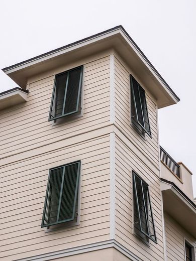 Corner of a light beige building with four open windows with dark green shutters.