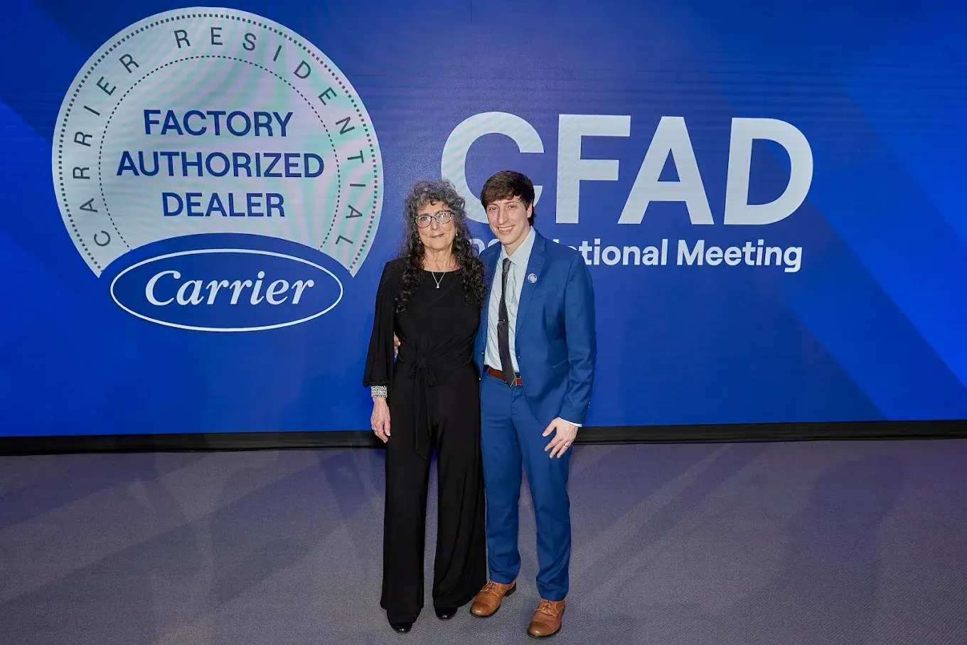 Two people posing in front of a blue CFAD Carrier conference backdrop