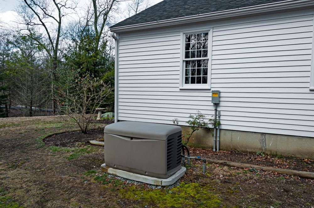 Off-white home generator on gravel bed next to a blue-sided house with windows and electrical boxes.