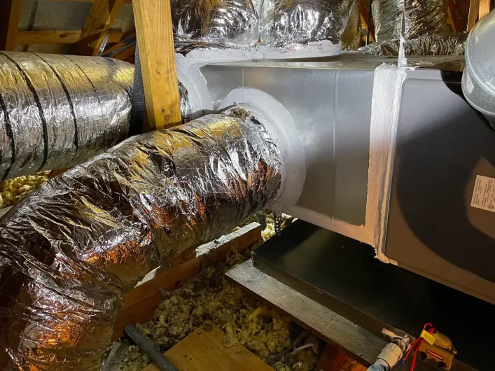 Insulated HVAC ductwork in a cramped attic or crawlspace, with silver foil-wrapped vents and a rectangular opening.