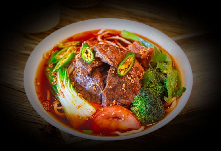 A bowl of spicy red soup with beef, noodles, bok choy, broccoli, and sliced green chilies