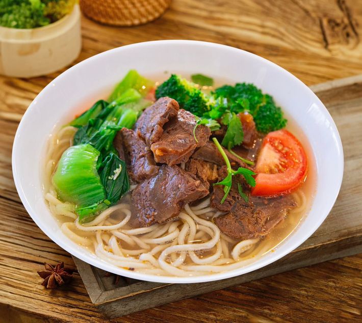 Bowl of noodle soup with beef, bok choy, broccoli, and tomato on a wooden table