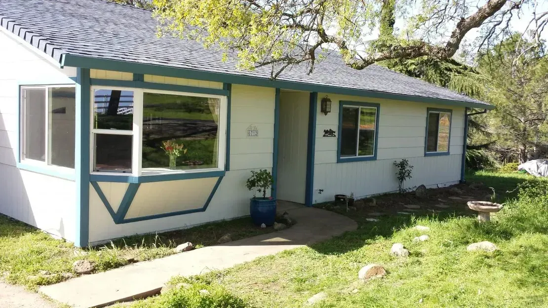 White house with blue trim, bay window, and path leading to the front door. Green grass and trees surround it.