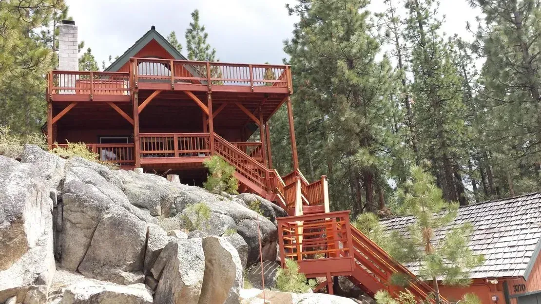 Red wooden cabin on a rocky hillside with stairs and a deck, surrounded by trees.