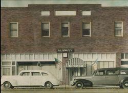 Vintage brick storefront with two parked cars in front and an awning over the entrance.
