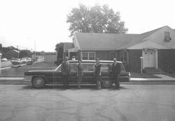 Three people stand beside a vintage car in front of a small building.