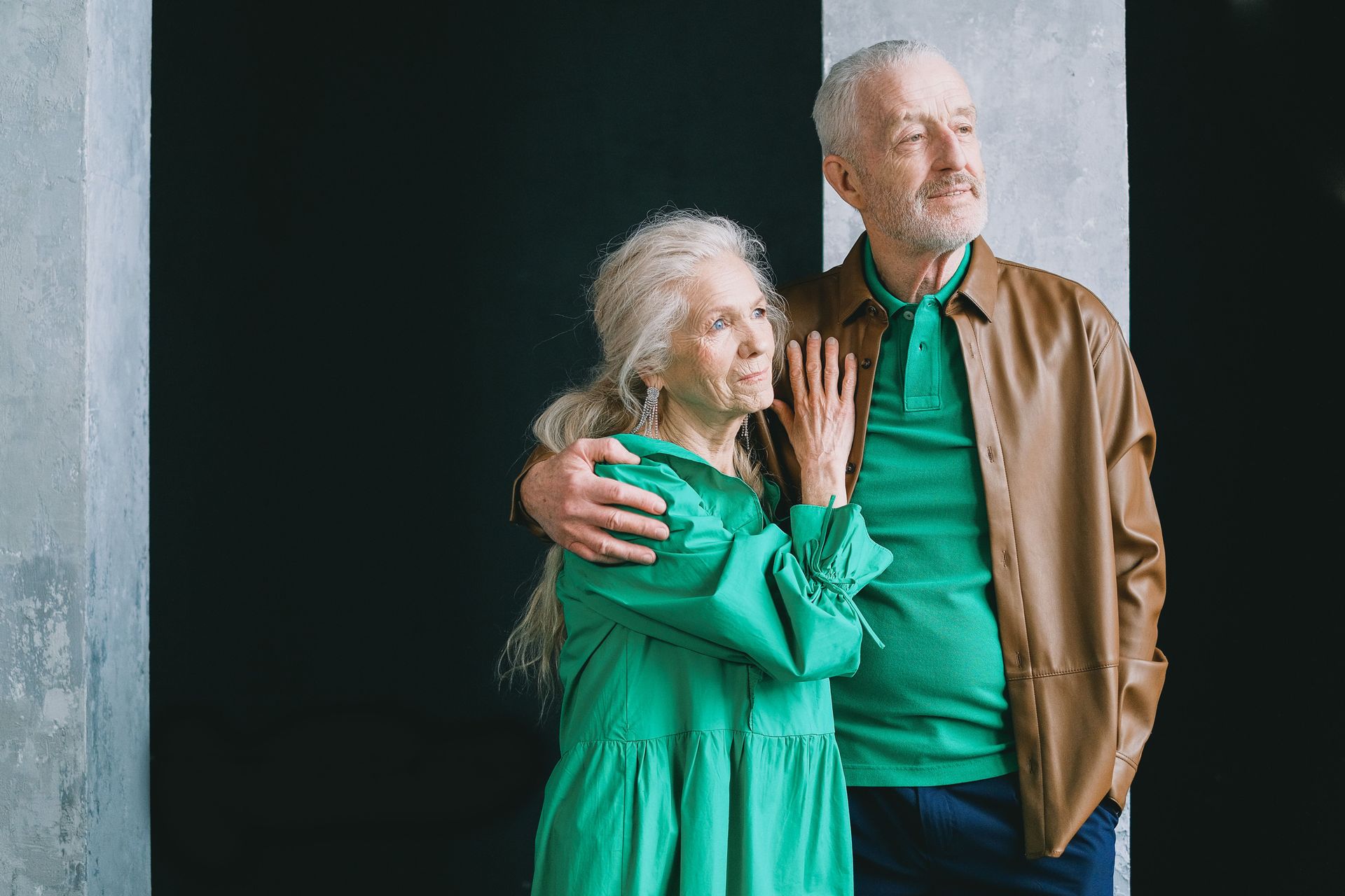 Two people standing close together by a window, smiling and embracing, one in a green dress and one in a tan jacket