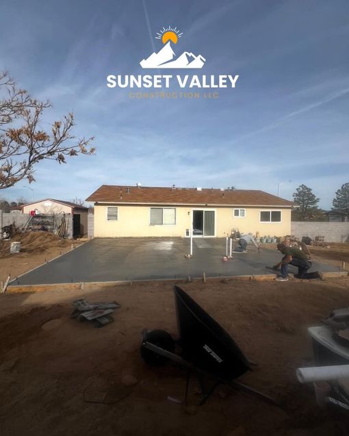 Concrete patio being poured near a beige house under a blue sky, with workers present.