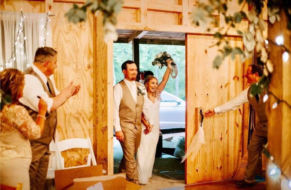 Newlyweds walk down a rustic barn aisle as guests applaud, bride holding bouquet overhead