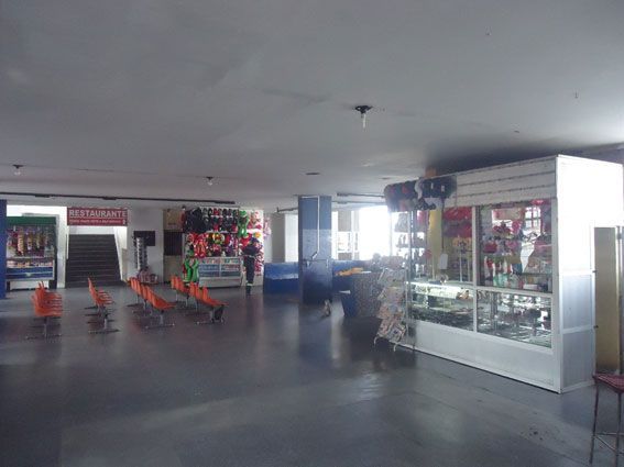 Interior vazio de um aeroporto com lojas e assentos. Paredes azuis e brancas, piso cinza.