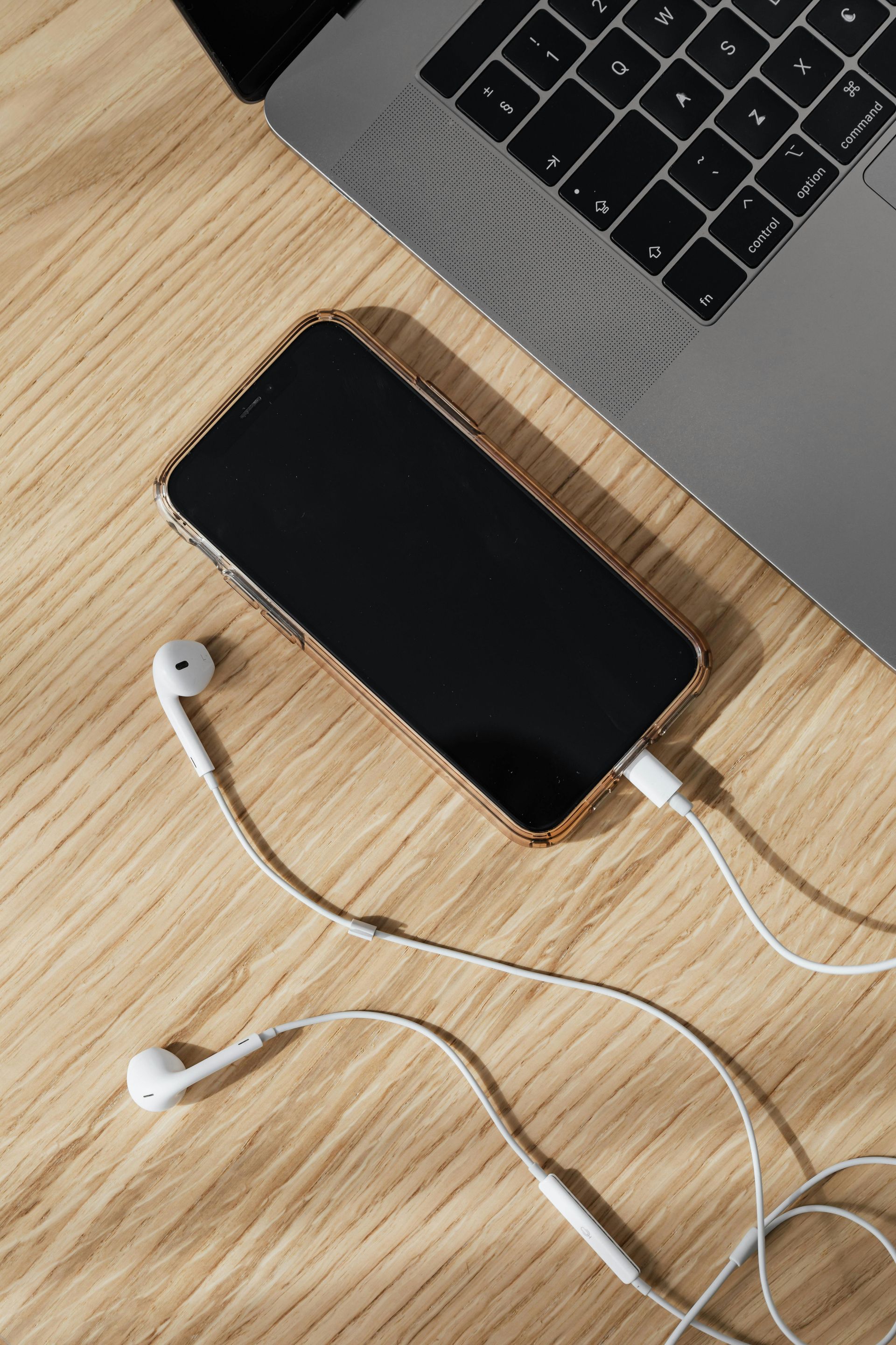 Smartphone com fones de ouvido com fio conectados, ao lado de um laptop sobre uma superfície de madeira.