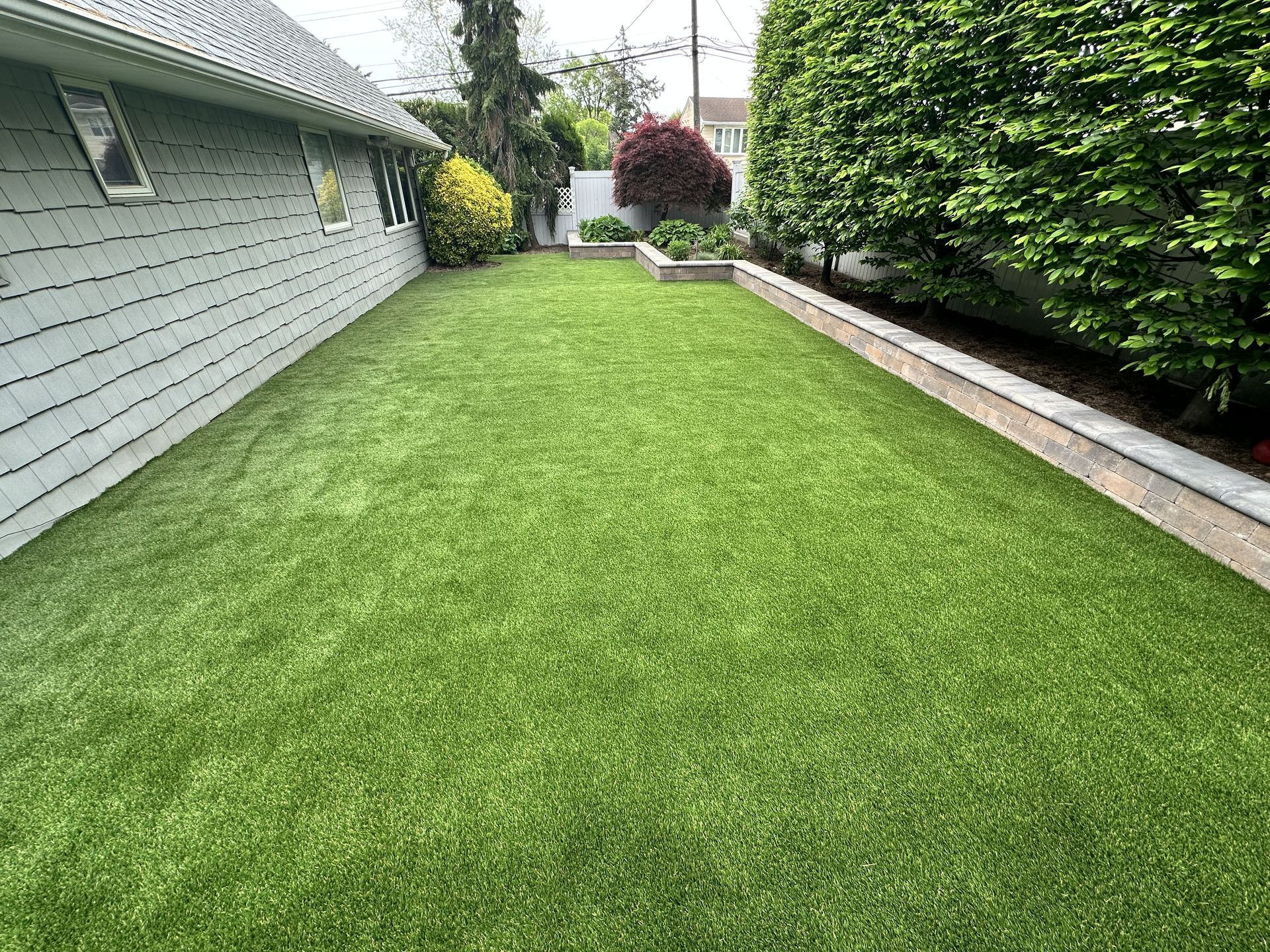 Green artificial lawn in a backyard, flanked by a house and shrubs, with a small garden bed.