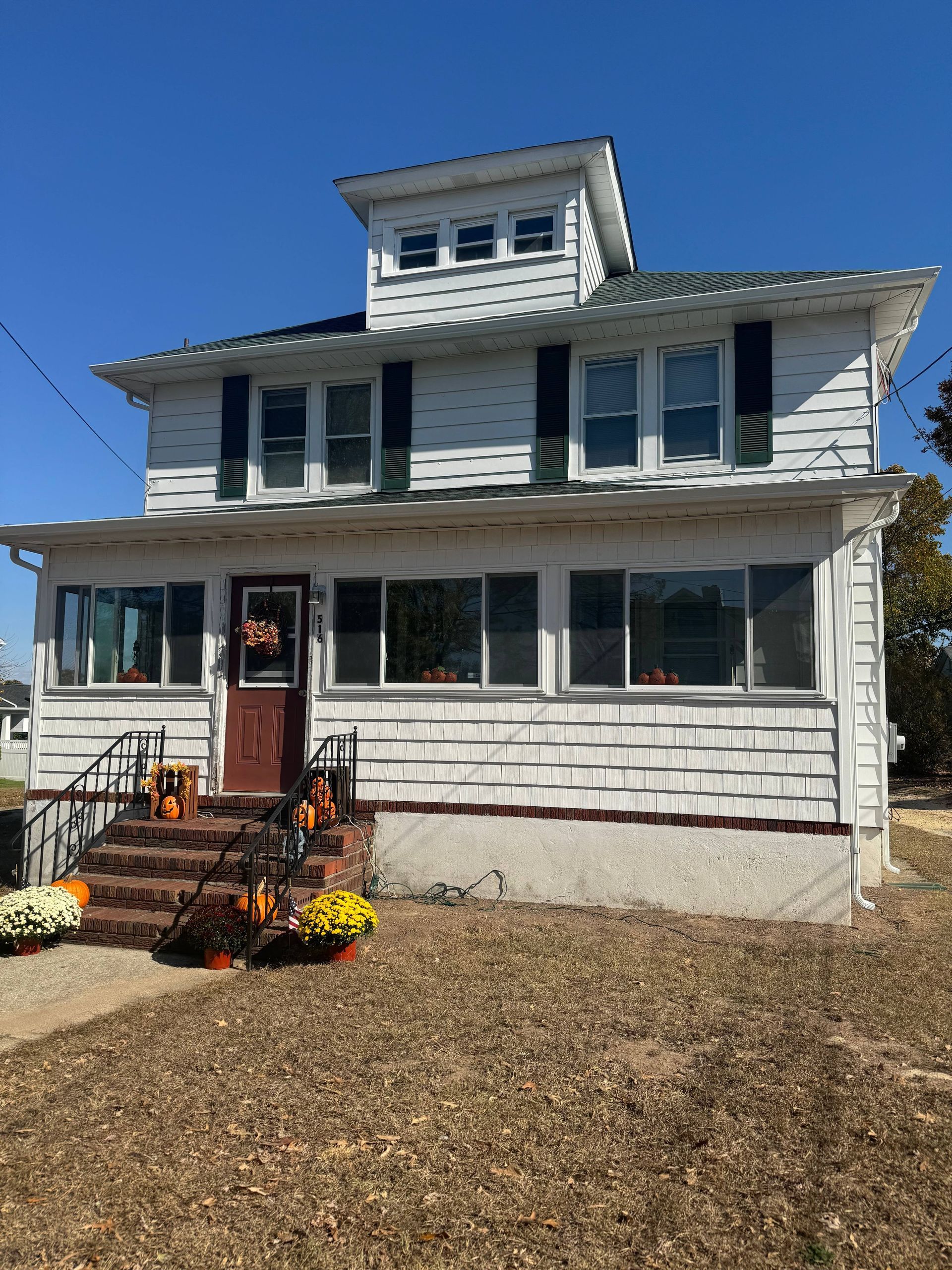 White two-story house with blue shutters, front porch, and autumn decorations under a clear blue sky