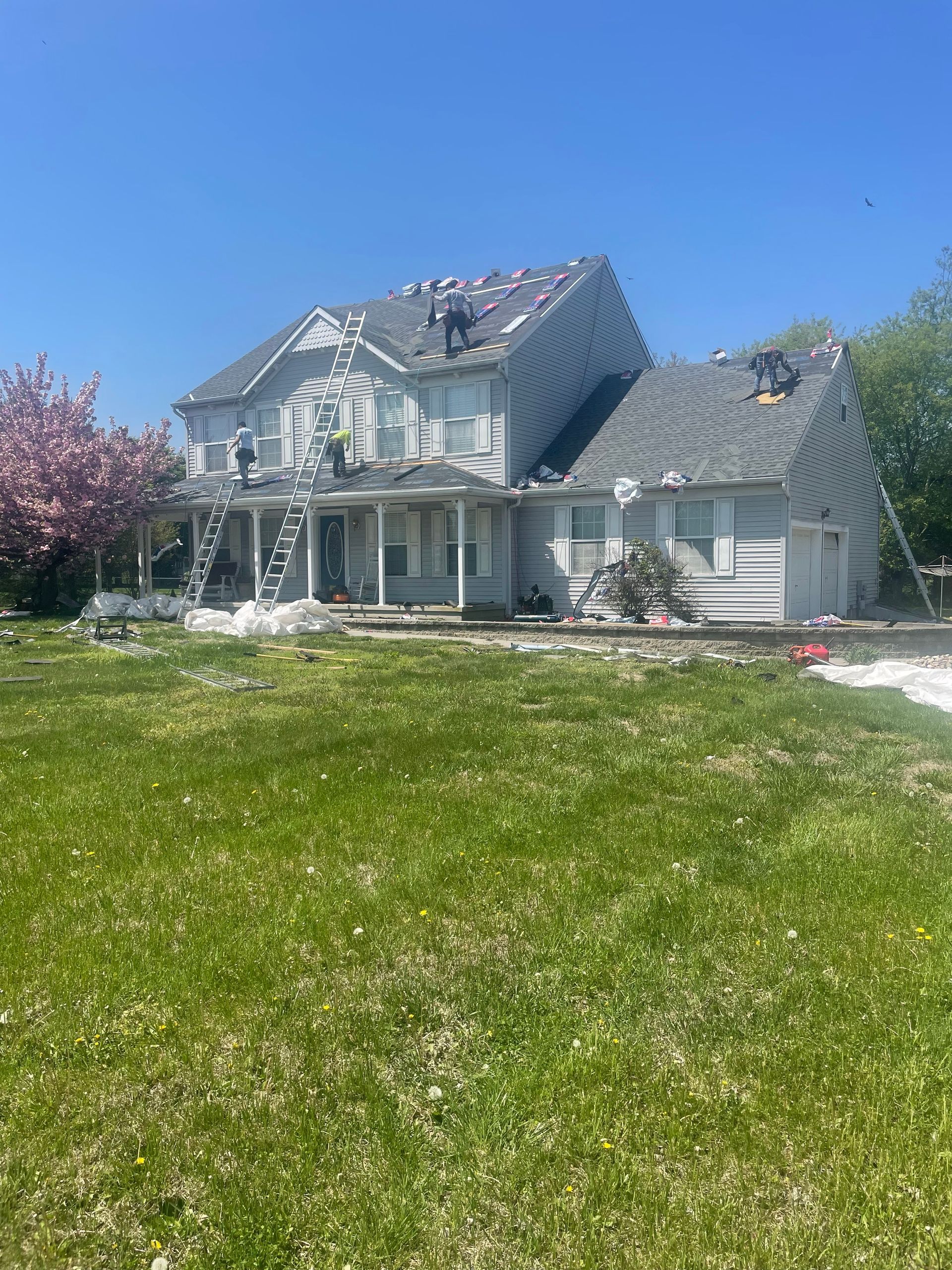 White two-story house with gray roof and porch, set in a sunny yard with green grass and pink flowering trees