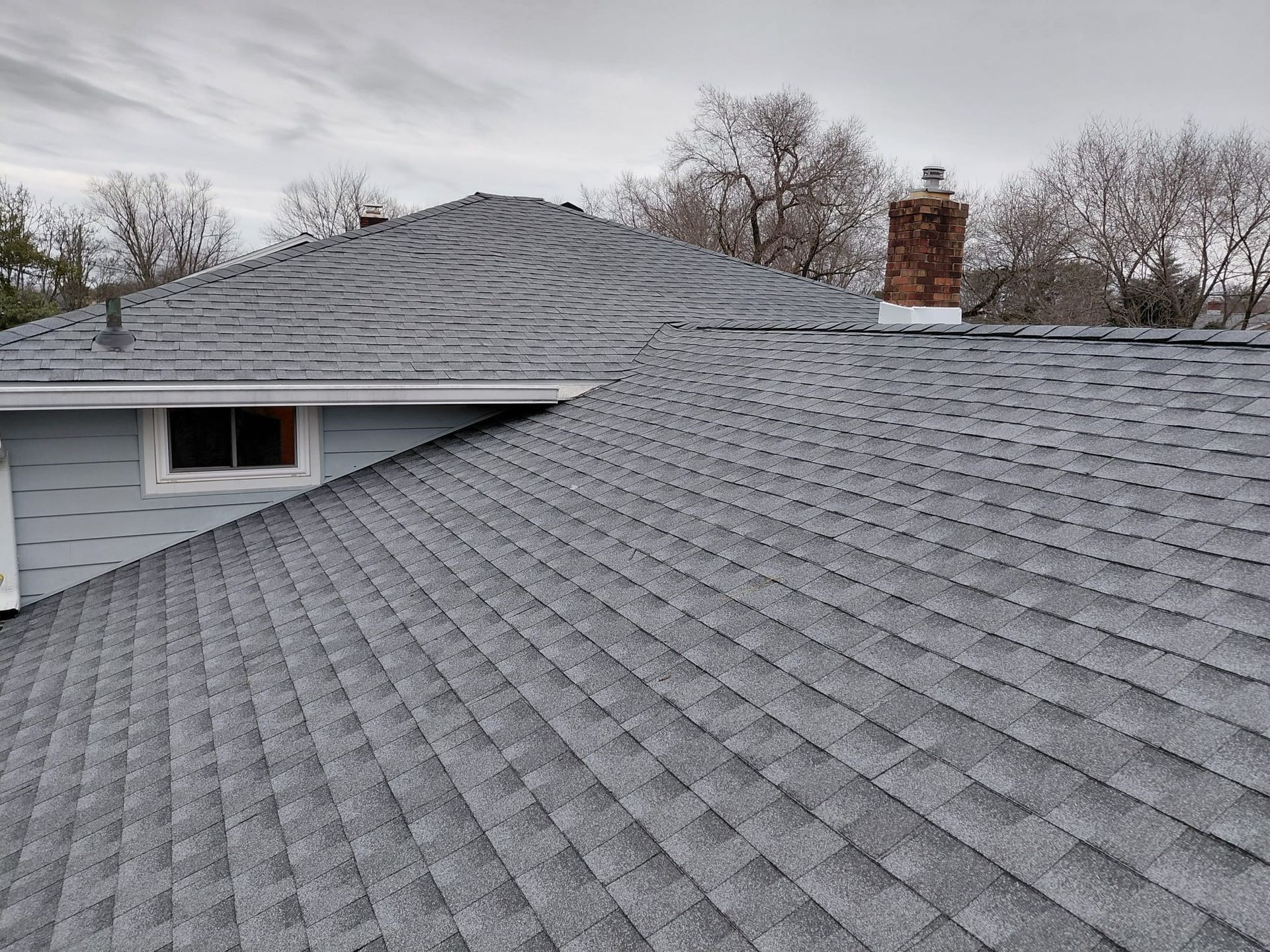 Gray shingled roof on a house with a brick chimney under an overcast sky.