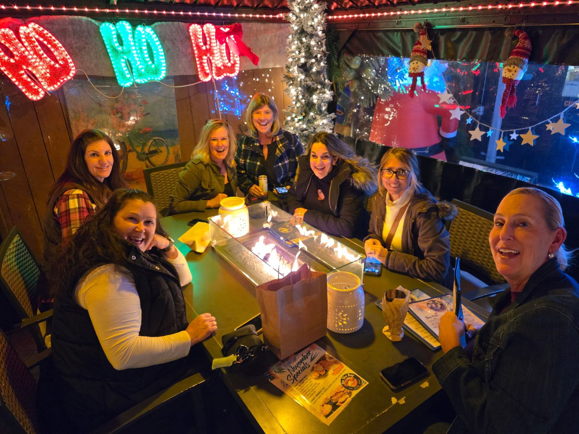 A group of women smiles around a table with a fire feature, festive decorations, and Christmas tree.