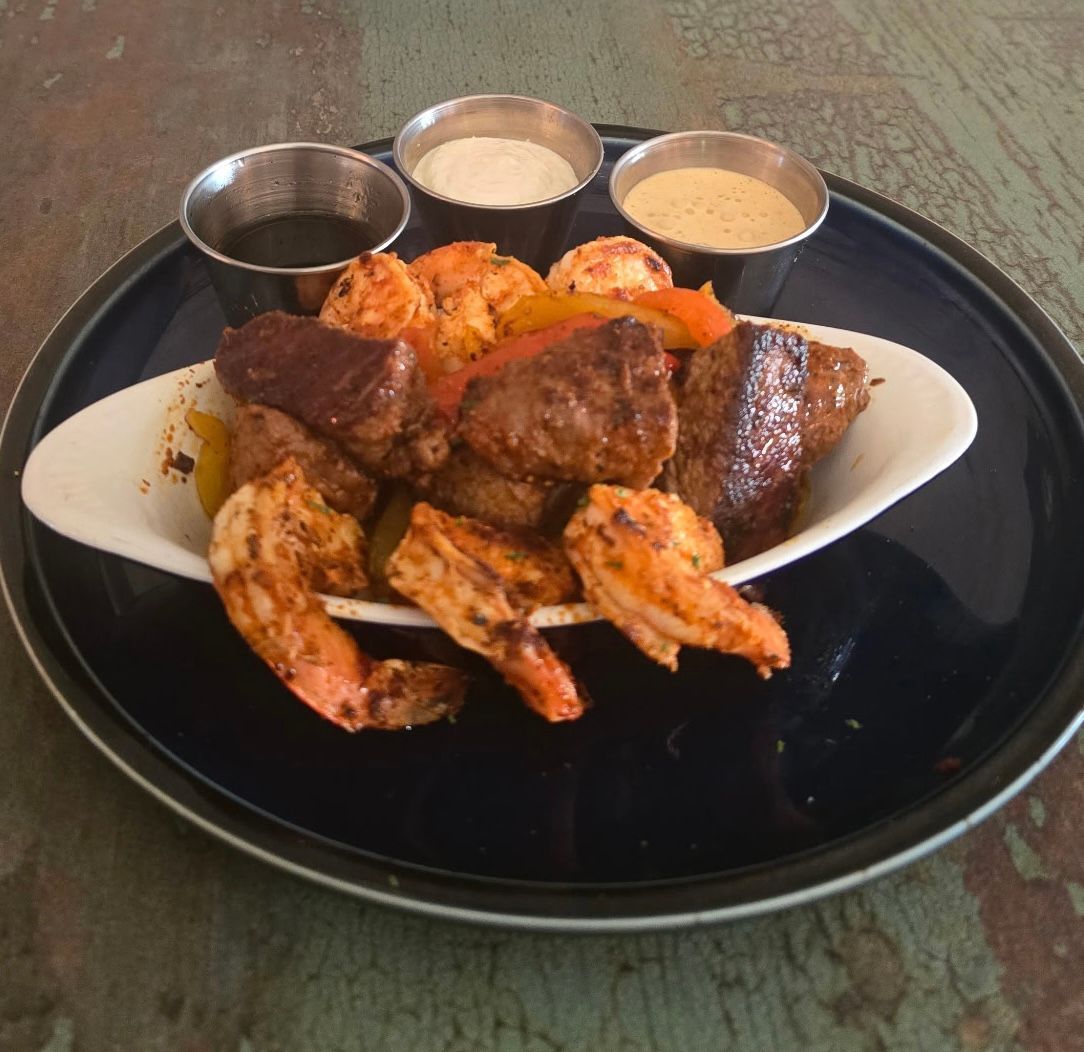 Grilled shrimp, steak, and peppers in a bowl with dipping sauces on a blue plate.