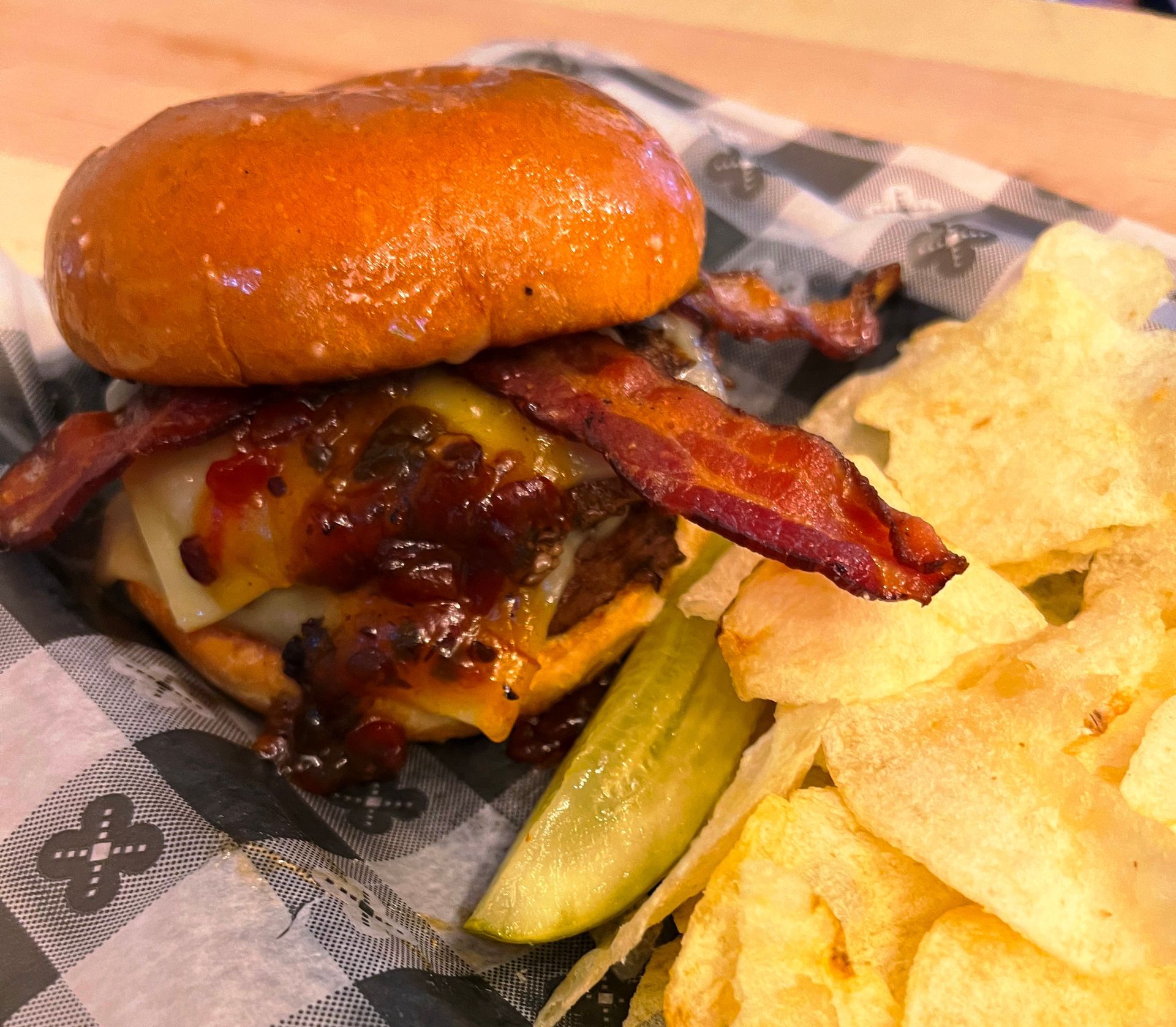 Burger with bacon, cheese, and caramelized onions, served with chips and a pickle.