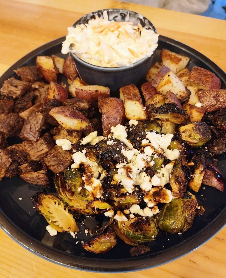Plate of roasted steak bites, potatoes, Brussels sprouts with feta, and coleslaw in a bowl.