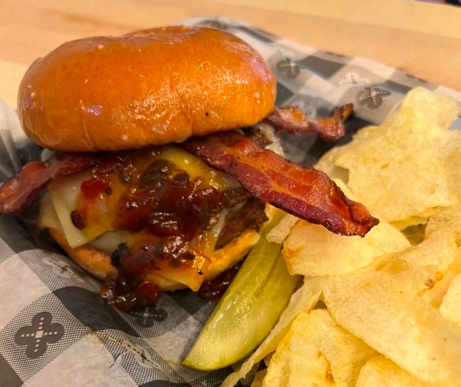 Burger with bacon, bacon jam, cheese, and sauce on a bun, served with chips and a pickle.