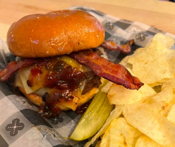 Burger with bacon, bacon jam, cheese, and sauce on a bun, served with chips and a pickle.