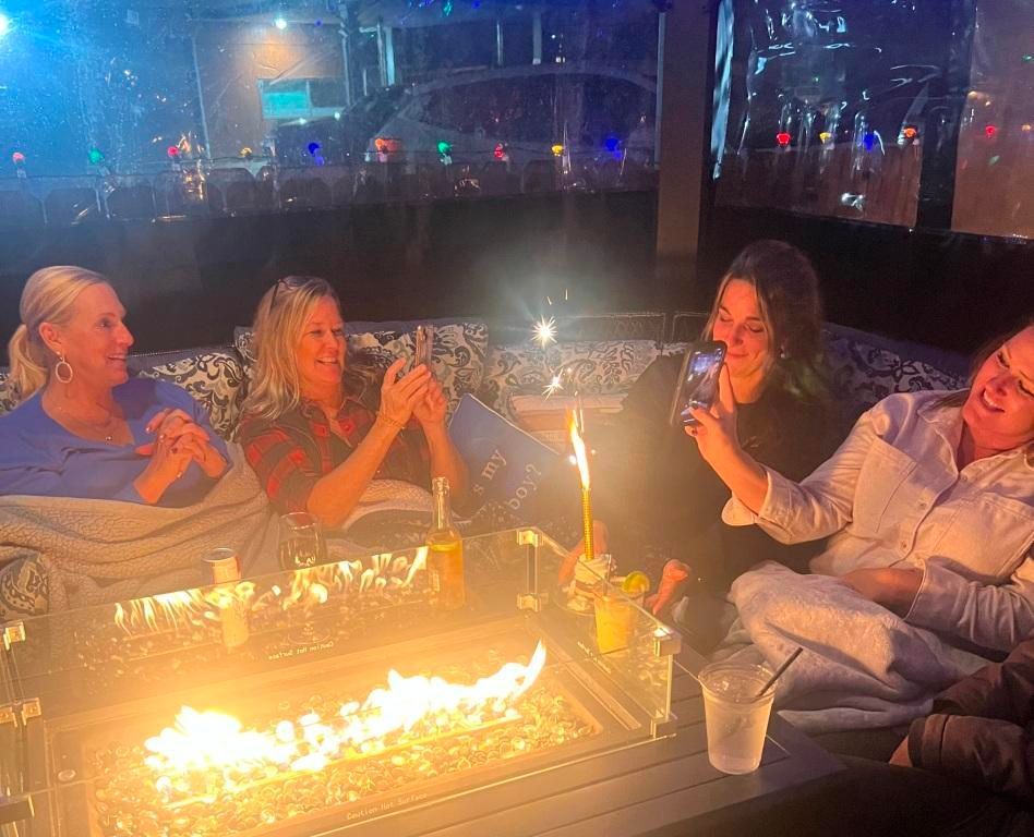 Four women by a fire pit, taking photos and laughing. Outdoor setting with lights.