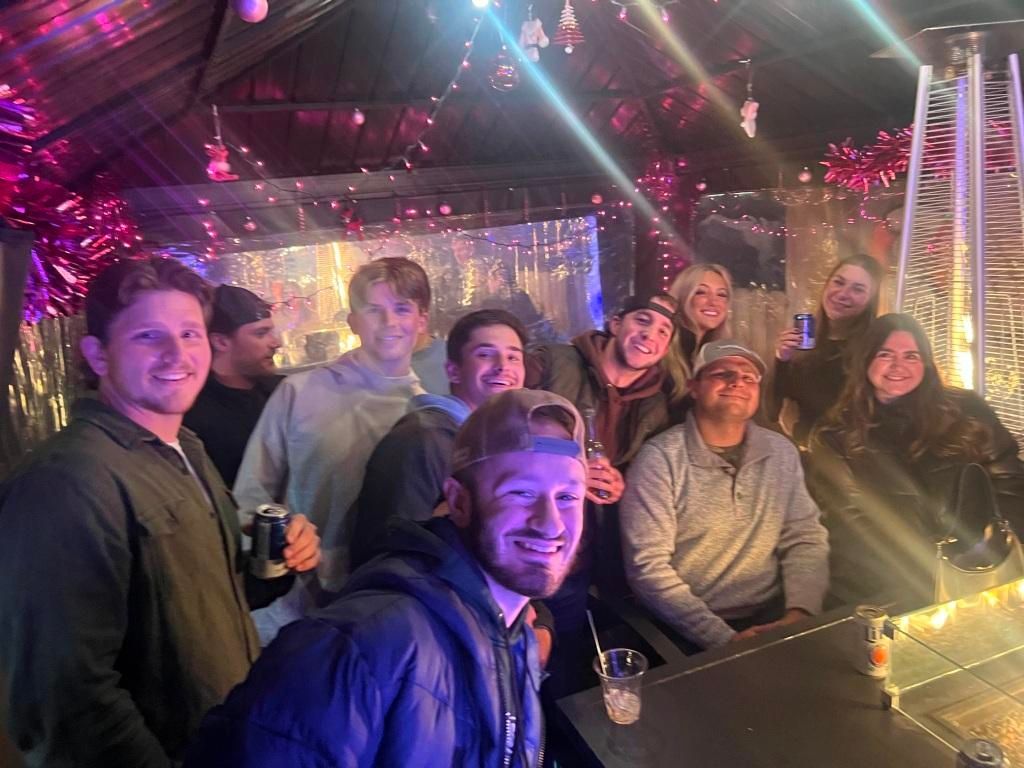 Group of friends smiling together, socializing outside.  Festive lights and heater visible.
