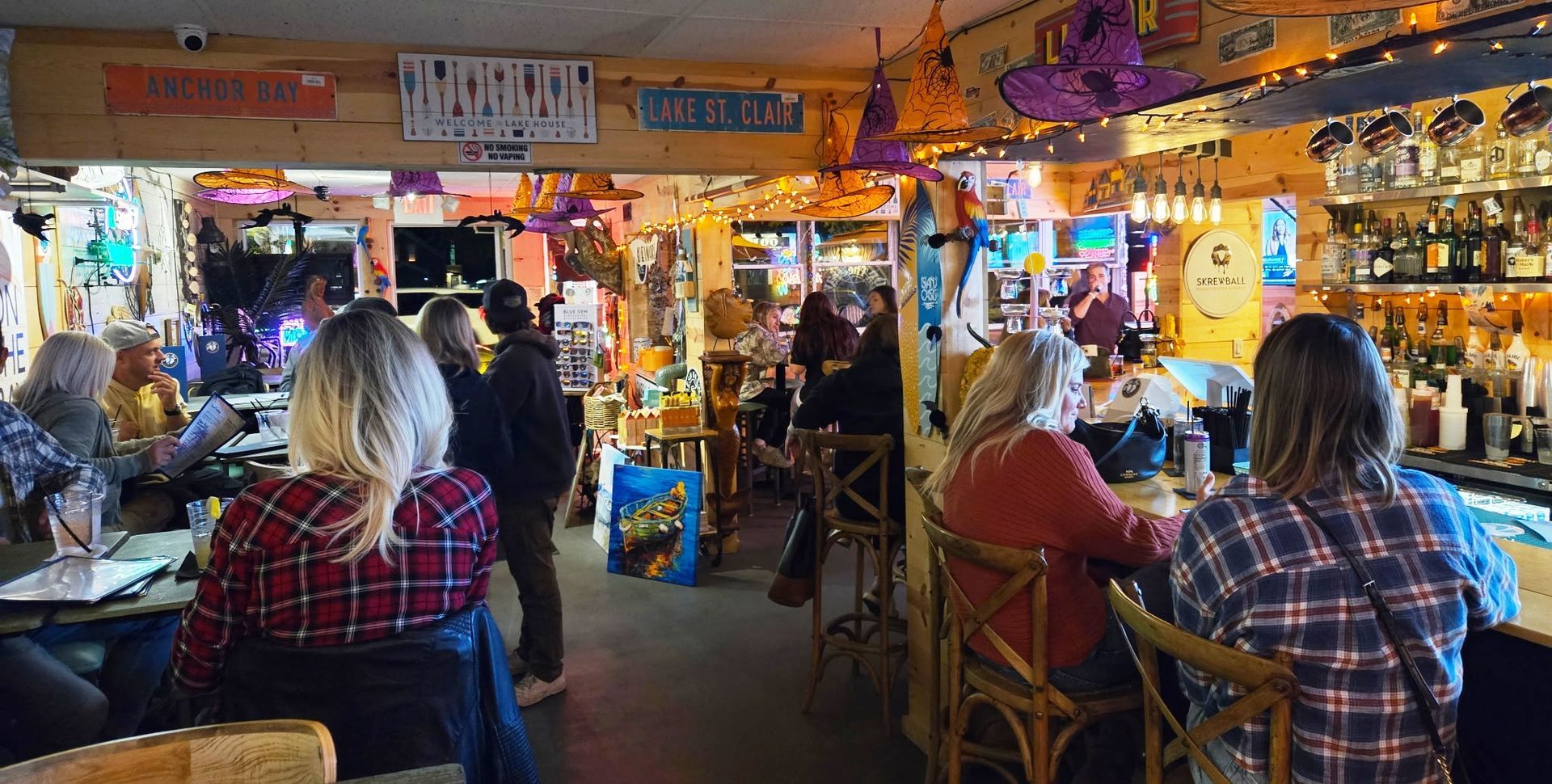 People gather in a warmly lit bar, some seated at the bar, others at tables. Decorated with festive lights and signs.