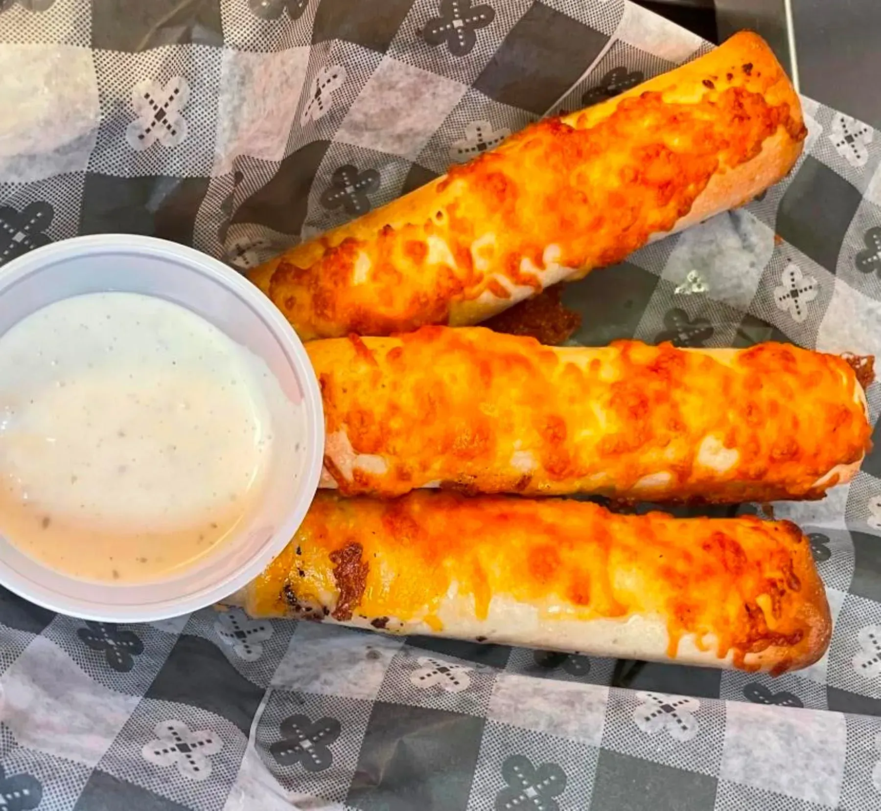 Three cheese breadsticks with dipping sauce on checkered paper.