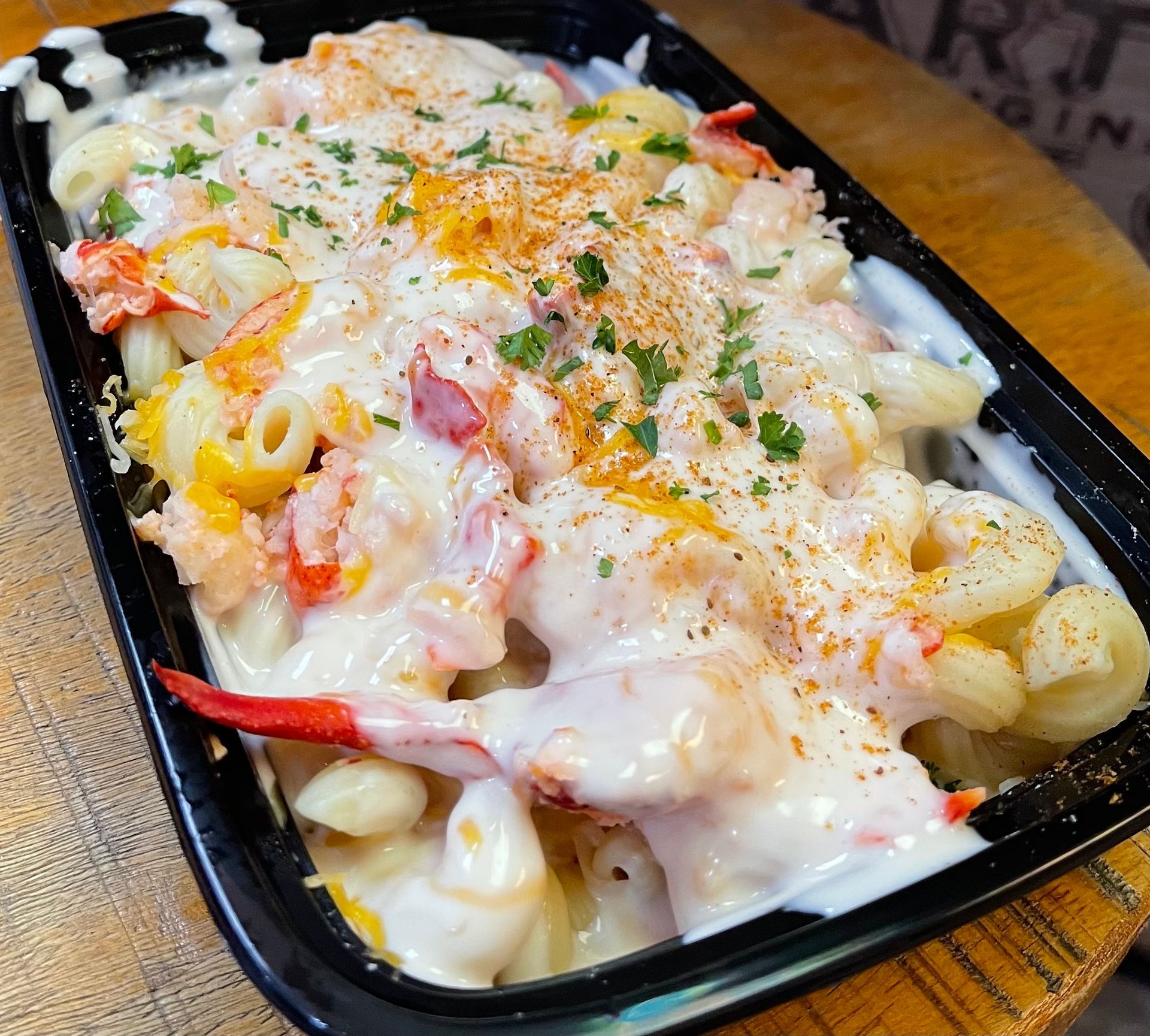 Lobster mac and cheese in a black container, creamy white sauce, topped with parsley and seasoning.