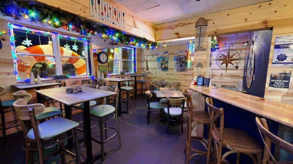 Interior of a nautical-themed bar with wooden walls, tables, and bar stools, decorated with marine art and garland.