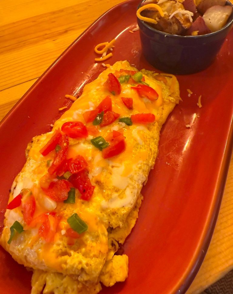 Omelet topped with tomatoes, green onions, and cheese on a red plate, served with a side of potatoes.