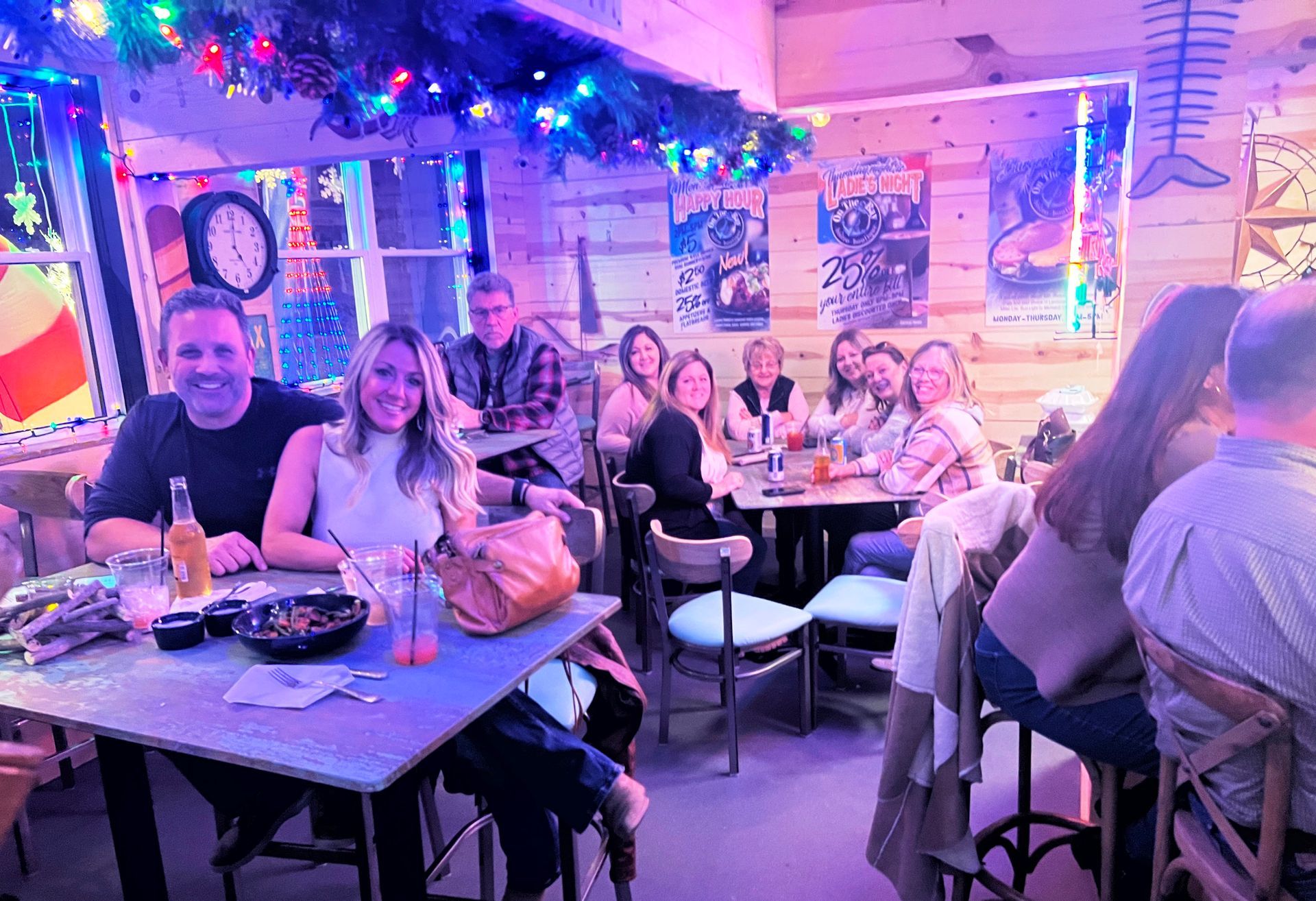 People at tables in a brightly lit restaurant, some with drinks and food. Party atmosphere with decorations.