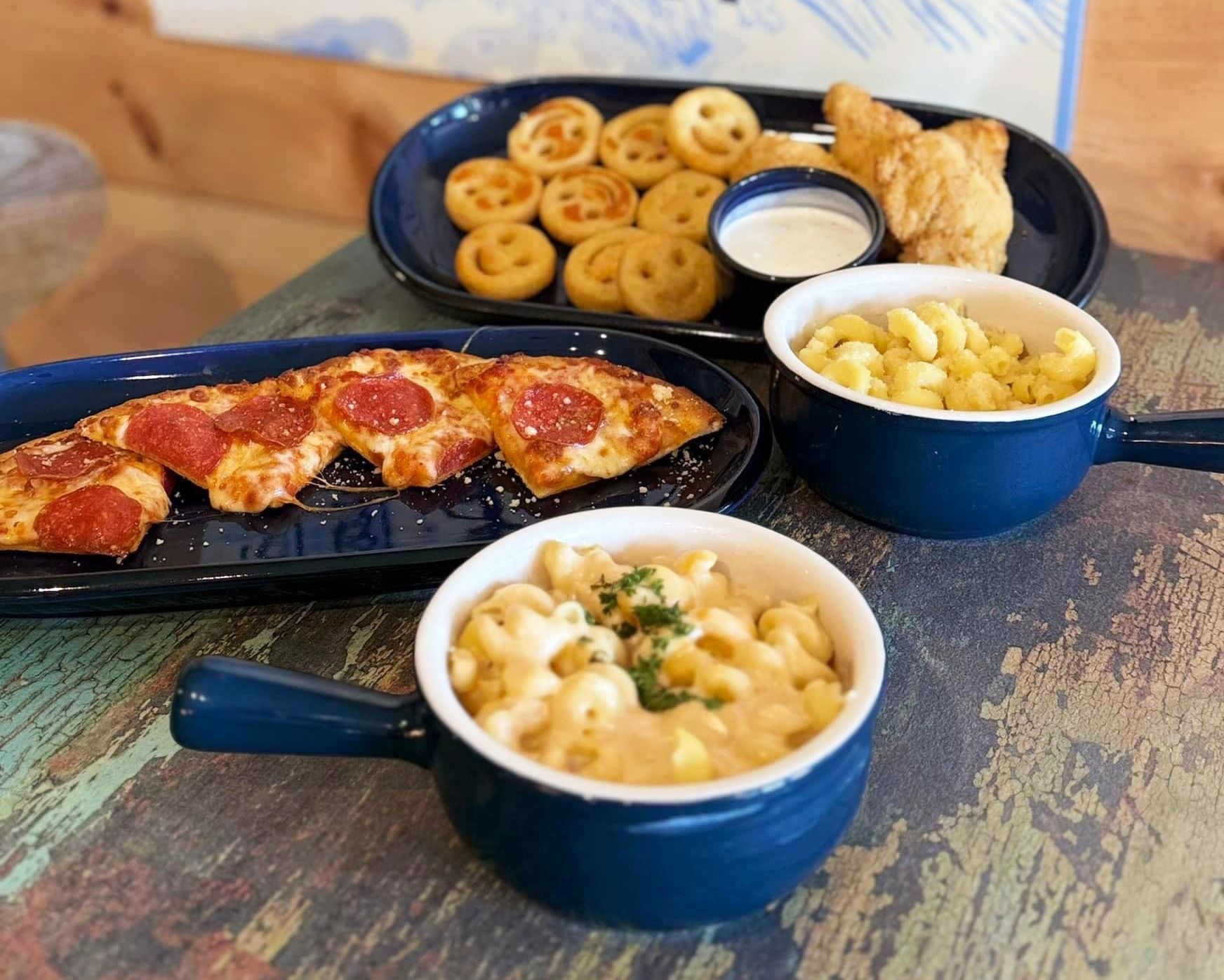 Platter of pizza slices, smiley fries, chicken nuggets, and mac and cheese in blue bowls on a wooden table.