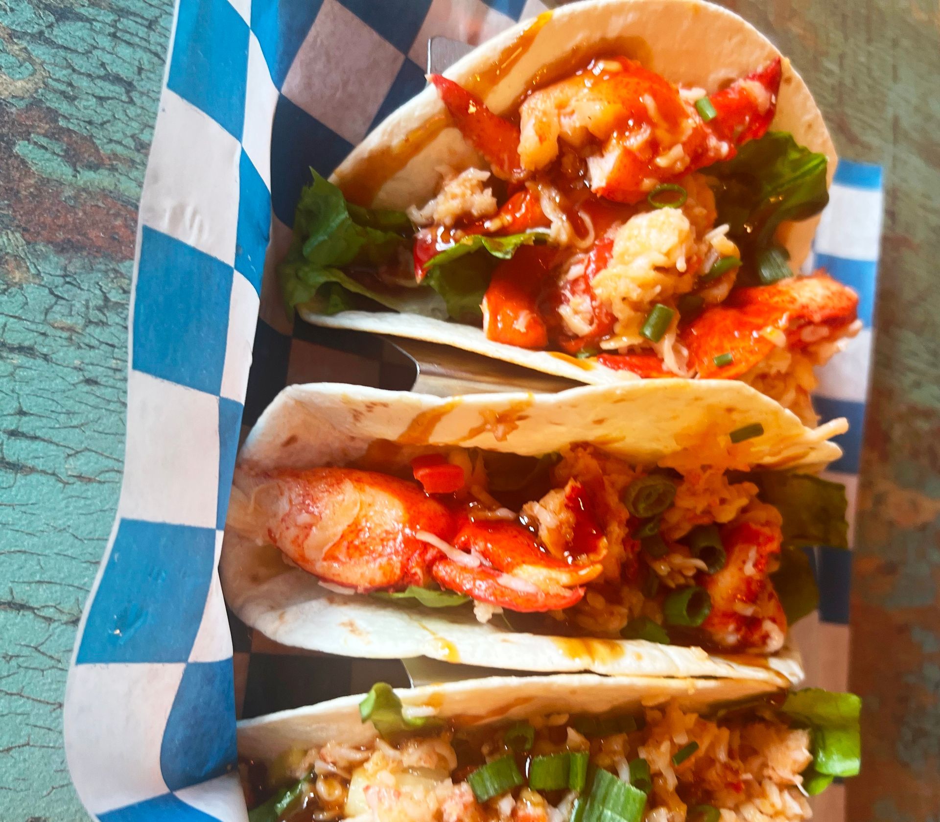 Three lobster tacos with green onions and sauce, served in a blue and white checkered paper holder.