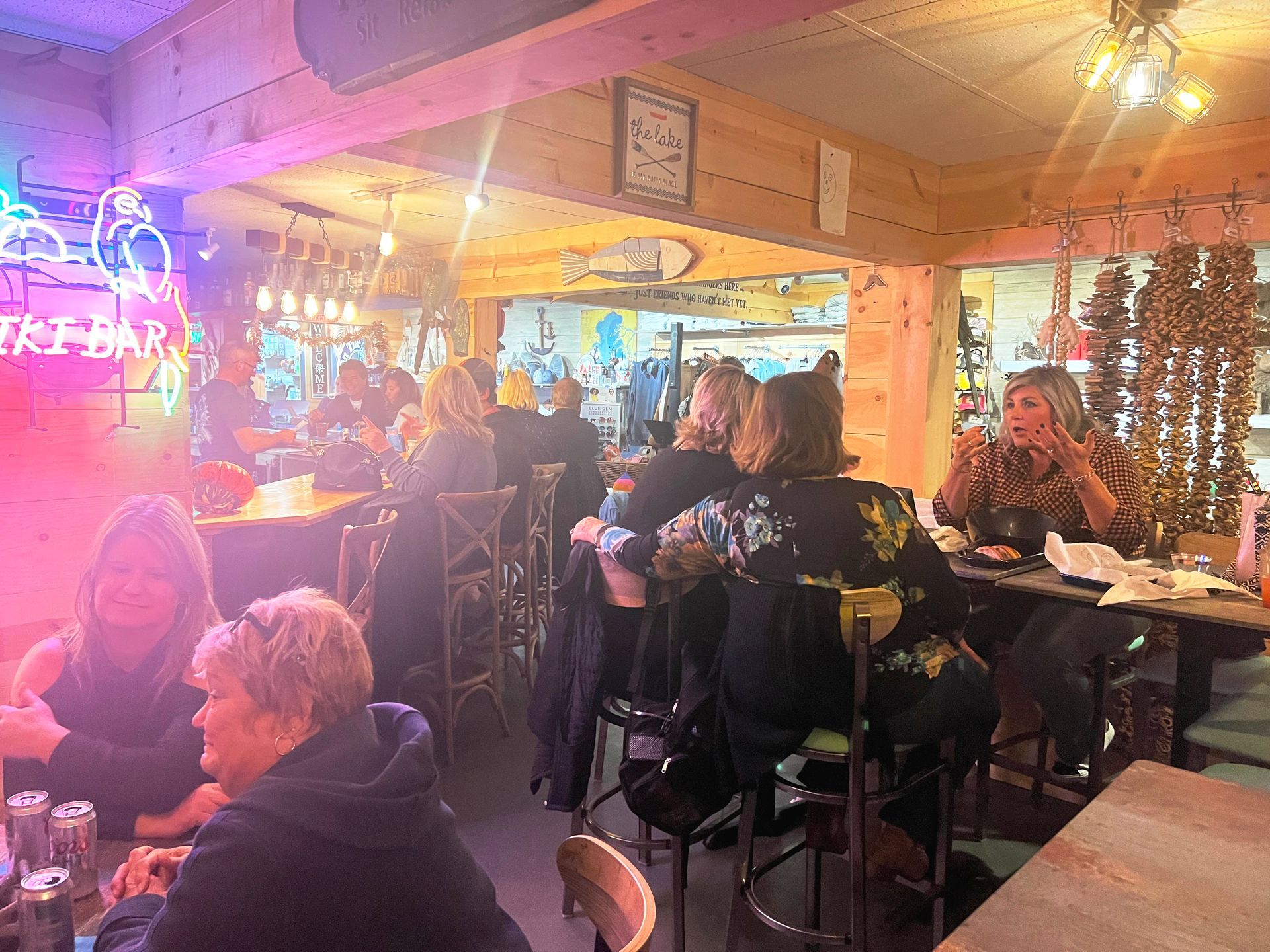 Interior of a bar with people seated at tables, socializing. Neon sign, warm lighting, wooden decor.